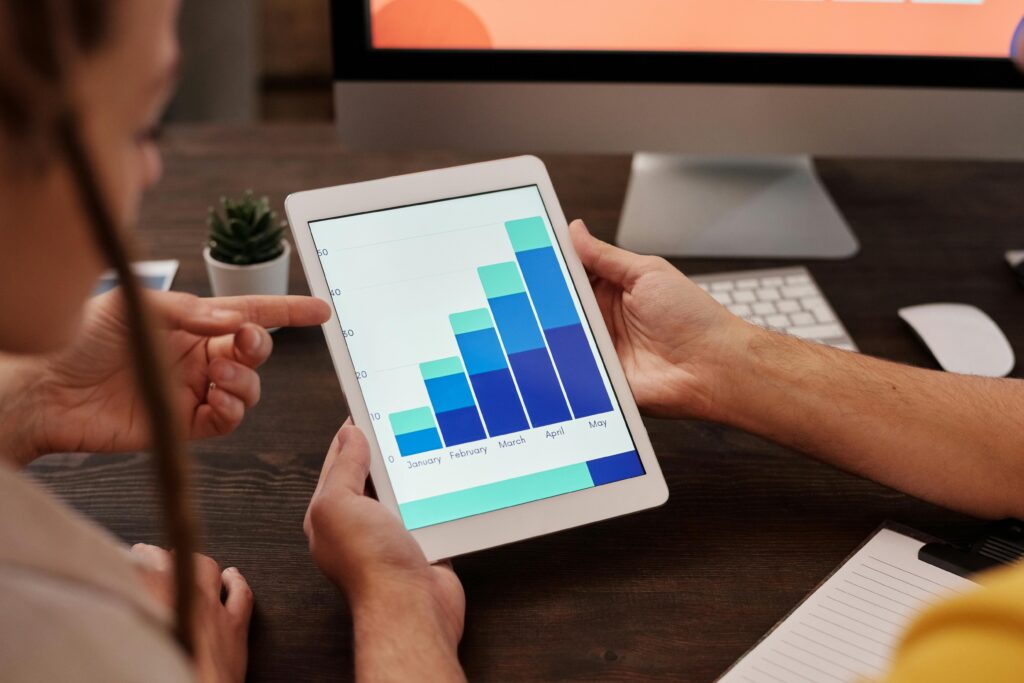 Google Analytics for SEO. Two people analyzing a bar chart on a tablet, showing business growth from January to May, with a desktop computer and notepad on the desk in the background