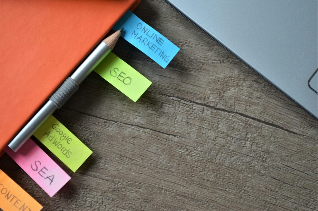Search Engine Optimization Close-up of a wooden desk with an orange notebook, colorful sticky notes labeled "ONLINE MARKETING," "SEO," "Google AdWords," "SEA," and "CONTENT," a pencil, and part of a laptop